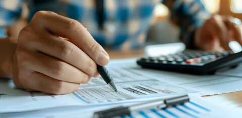 Hand holding a pen over printed financial documents with graphs, analyzing expenses and calculating figures.