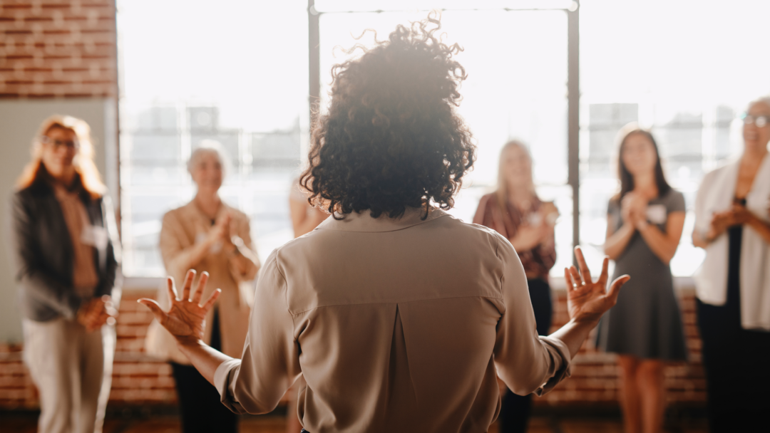 Back view of a woman speaking to a group of people in a bright room, with several women standing and clapping.