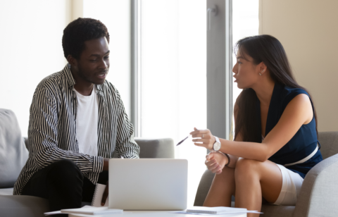 Two people sitting in a modern office setting, having a focused conversation in front of a laptop.
