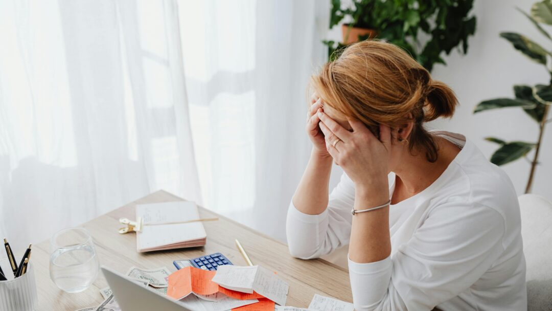 Woman sitting at a desk with her head in her hands, surrounded by bills and receipts, appearing stressed.