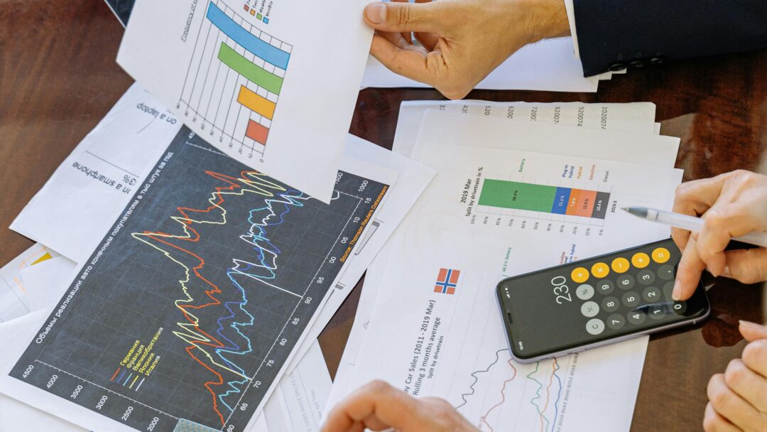 Two professionals review printed charts and graphs at a meeting table, using a calculator and laptop to analyze financial or marketing data.