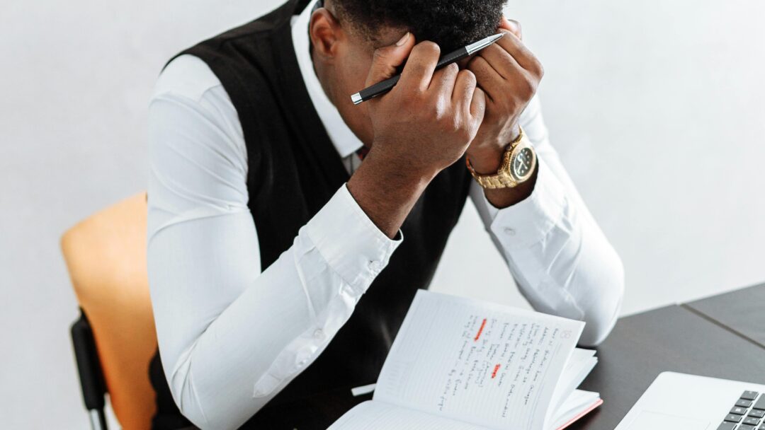 Man sitting at a desk with his head in his hands, surrounded by a laptop, notebook, and hourglass.